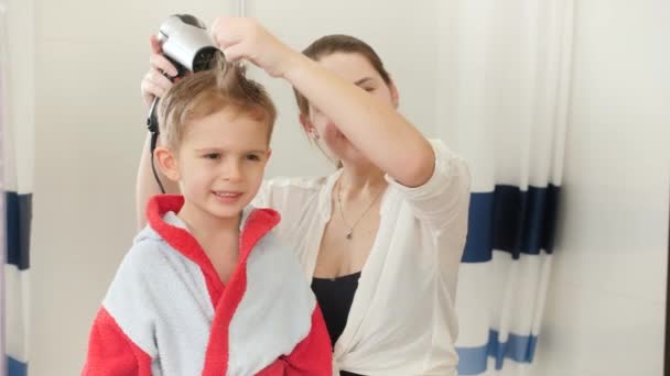 Маленький улыбающийся мальчик в халате и мама сушит его волосы, стоя перед зеркалом в ванной. Концепция детской гигиены и здравоохранения на дому. Забота о родителях и детях — стоковое видео