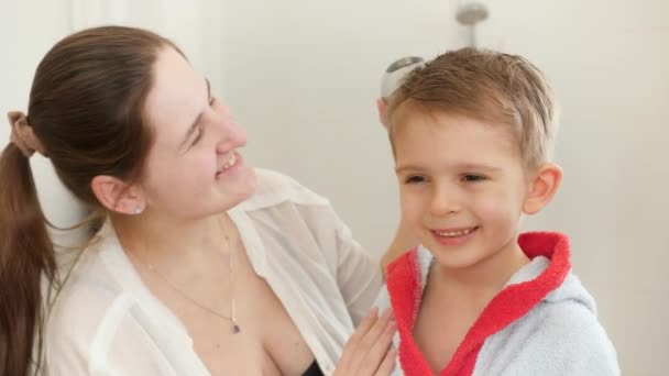 Mignon petit garçon en peignoir regardant dans le miroir tandis que la mère sèche ses cheveux avec sèche-cheveux dans la salle de bain. Concept d'hygiène infantile et de soins de santé à domicile. Parents bienveillants et enfants à la maison — Video