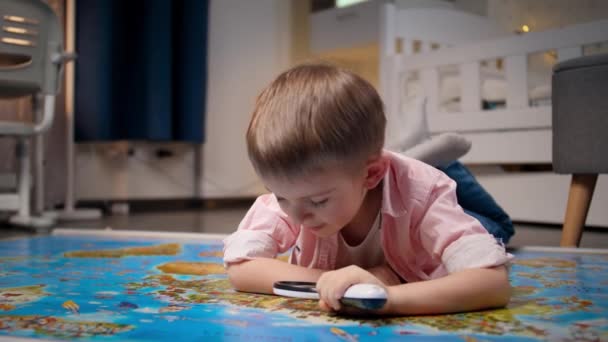Маленький усміхнений хлопчик лежить на підлозі в кабінеті шланга і досліджує велику карту світу через збільшувальне скло. Концепція подорожей, туризму та виховання дітей. Дослідження та відкриття дітей — стокове відео
