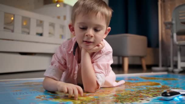 Милий усміхнений хлопчик лежить на підлозі і ходить по країнах з пальцями на великій карті світу. Концепція подорожей, туризму та виховання дітей. Дослідження та відкриття дітей . — стокове відео