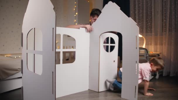 Szczęśliwy uśmiechnięty chłopczyk bawiący się w swoim pokoju, podczas gdy ojciec montuje domek z kartonu. Rodzina dobrze się bawi. Dzieci pomagające rodzicom. — Wideo stockowe
