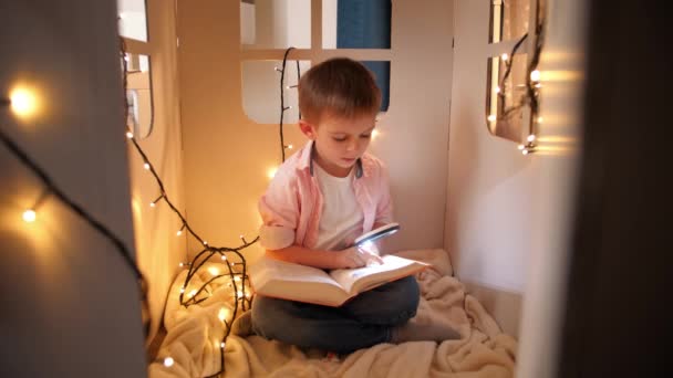 Милый маленький мальчик сидит в своем игрушечном домике и читает книгу с лупой и факелом. Концепция детского образования и чтения в темной комнате — стоковое видео
