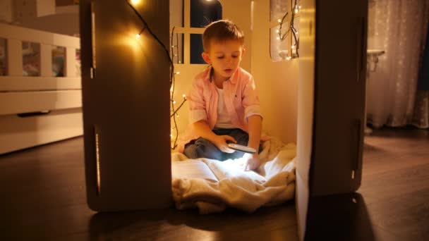 Dolly skott av liten pojke med ficklampa och förstoringsglas för att läsa bok på natten. Begreppet barnuppfostran och läsning i mörkrum — Stockvideo