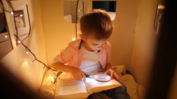 Вид через вікно іграшкового будинку на маленькому хлопчику читає книжку вночі. Концепція виховання дітей та читання в темній кімнаті — стокове відео