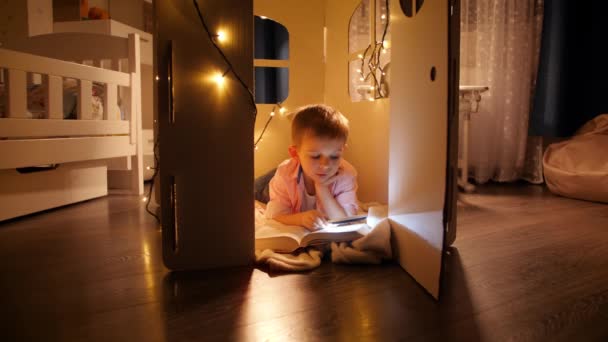 Joyeux garçon souriant couché sur le sol à la maison de jouets hos ou tente et la lecture avec une lampe de poche la nuit. Concept d'éducation et de lecture des enfants en chambre noire — Video