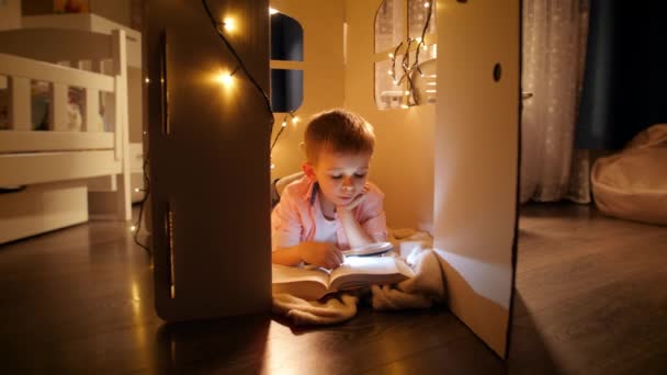 Паннінг маленького розумного хлопчика, який читає велику книгу вночі з ліхтариком. Концепція виховання дітей та читання в темній кімнаті — стокове відео