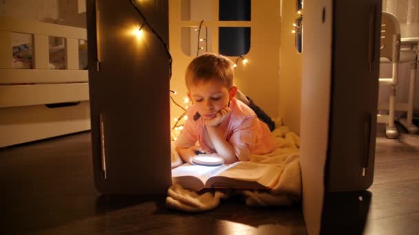 Dolly Shot von Jungen, die Buch lesen, während sie nachts in Spielzeughaus oder Zelt liegen. Konzept der Kindererziehung und des Lesens im dunklen Raum — Stockvideo