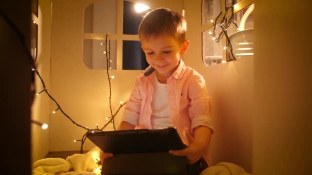Портрет щасливого сліпучого хлопчика, який використовує планшетний комп'ютер і переглядає Інтернет, граючи у своєму іграшковому картонному будинку або наметі вночі. Концепція виховання дітей та навчання вночі — стокове відео