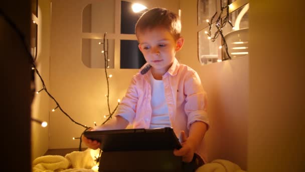 Портрет маленького мальчика, смотрящего мультики на планшетном компьютере ночью, сидящего в маленьком игрушечном домике. Концепция детского образования и обучения в ночное время — стоковое видео