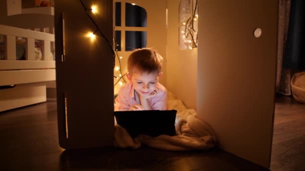 Фотография улыбающегося мальчика с планшетным компьютером, лежащим на полу в палатке или в картонном домике. Концепция детского образования и обучения в ночное время — стоковое видео