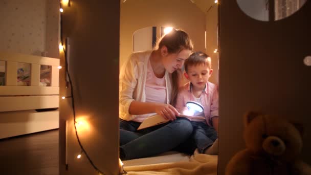 Szczęśliwy uśmiechnięty chłopiec z młodą matką siedzącą w namiocie lub kartonowym domu zabawki i czytającą książkę z latarką w nocy. Pojęcie wychowania dzieci i rodziny spędzających razem czas w nocy — Wideo stockowe