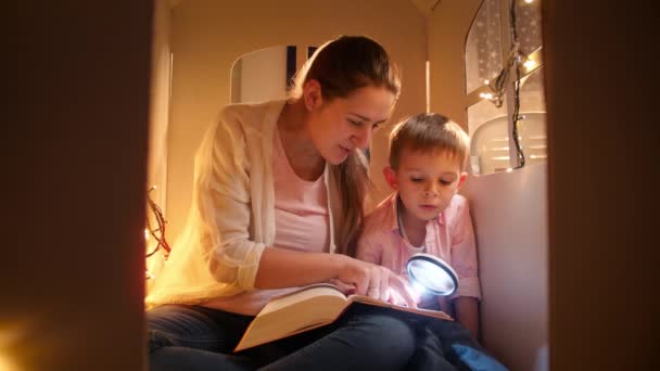 Портрет улыбающейся молодой матери с маленьким мальчиком, читающим ночью книгу во время игры в игрушечном домике. Концепция детского образования и семейного времяпровождения вместе ночью — стоковое видео