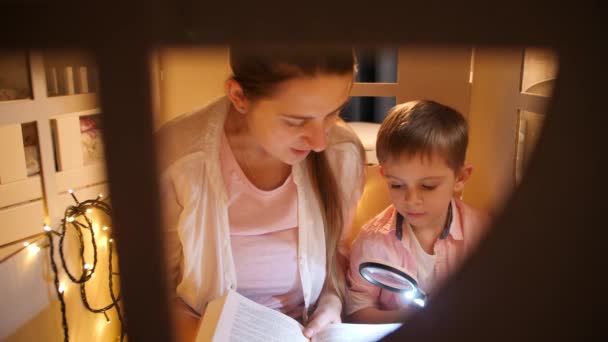 Retrato de menino bonito com mohter jovem lendo grande livro na barraca ou casa de brinquedo antes de ir dormir. Conceito de educação infantil e família tendo tempo juntos à noite — Vídeo de Stock