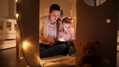 Çadırda ya da oyuncak evde oynayan ve tablet bilgisayar kullanan küçük, gülümseyen bir çocuk. Çocuk eğitimi ve aile kavramının geceleri birlikte vakit geçirmesi