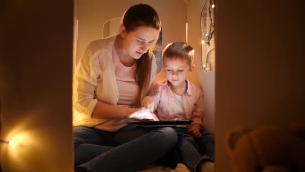 Oyuncak evinde ya da çadırında oğluyla oturup tablet bilgisayar kullanan gülümseyen bir anne resmi. Çocuk eğitimi ve aile kavramının geceleri birlikte vakit geçirmesi — Stok video