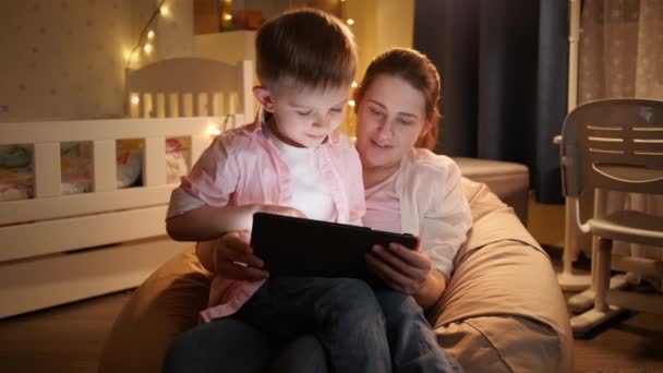 Porträt eines kleinen Jungen, der auf dem Schoß seiner Mutter sitzt und sich nachts Cartoons auf dem Tablet-Computer ansieht. Konzept von Kindererziehung und Familie, die nachts Zeit miteinander verbringen — Stockvideo