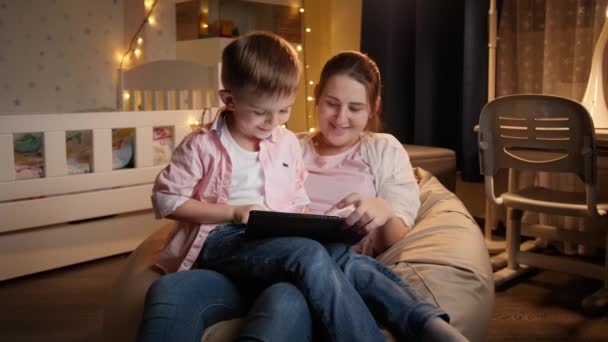 Mãe sorridente com filho pequeno sentado em saco de feijão à noite e cantar tablet computador. Conceito de educação infantil e família tendo tempo juntos à noite — Vídeo de Stock