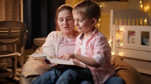 Милый маленький мальчик, сидящий с мамой по ночам и читающий сказки на ночь. Концепция детского образования и семейного времяпровождения вместе ночью — стоковое видео