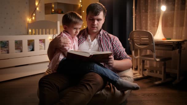 Junger Vater sitzt mit seinem kleinen Sohn im Schlafzimmer und liest Märchen. Konzept von Kindererziehung und Familie, die nachts Zeit miteinander verbringen — Stockvideo