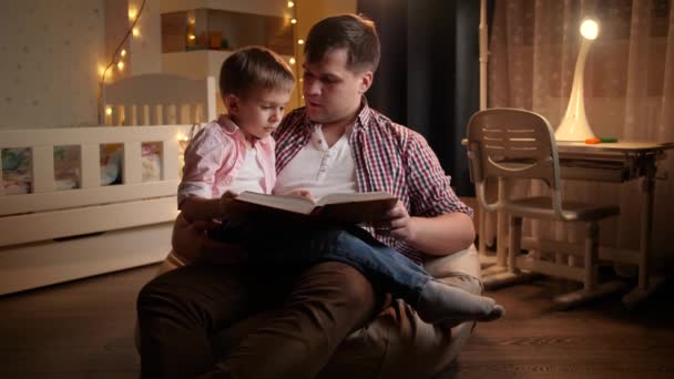Улыбающийся мальчик слушает сказку на ночь, которую ему читает отец. Концепция детского образования и семейного времяпровождения вместе ночью — стоковое видео