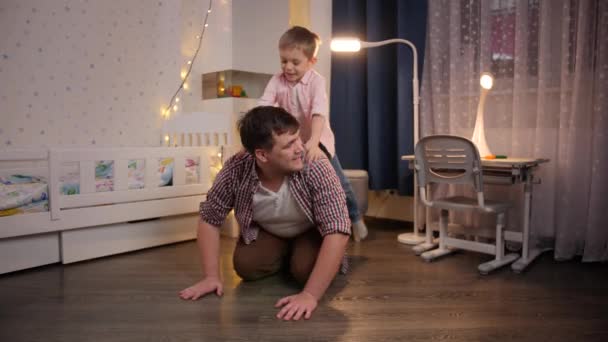 Un niño sonriente saltando sobre su padre y montando cerdito en el dormitorio. Concepto de niño jugando con los padres y la familia pasar tiempo juntos en la noche — Vídeos de Stock