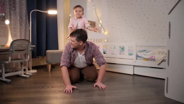 Feliz padre sonriente con su pequeño hijo jugando y montando cerdito en el dormitorio. Concepto de niño jugando con los padres y la familia pasar tiempo juntos en la noche — Vídeos de Stock