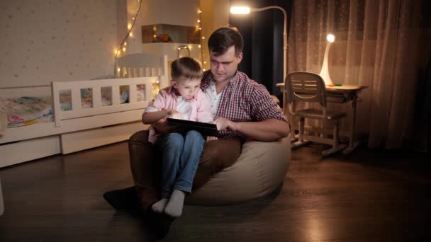 Szczęśliwy chłopak śmiejący się z młodym ojcem siedzącym w nocy w sypialni i grającym w gry na tablecie. Pojęcie wychowania dzieci i rodziny spędzających razem czas w nocy. — Wideo stockowe