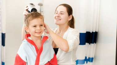 Banyoda yıkandıktan sonra saçlarını kurutmak için saç kurutma makinesi kullanan genç bir annenin portresi. Evde çocuk hijyeni ve sağlık hizmetleri kavramı. Evde ebeveynlere ve çocuklara bakmak.