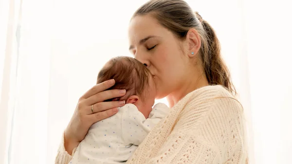 Closeup portrait of young loving mother kissing her little newborn baby at window — Stock Photo, Image