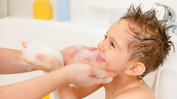 Retrato de un niño sonriente alegre con el pelo mojado lavándose en el baño con una madre joven y cariñosa. Concepto de higiene infantil y atención sanitaria en el hogar. Familia divirtiéndose y jugando. — Foto de Stock