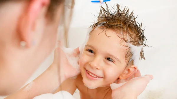 Mladá starostlivá matka myje svému synovi vlasy šamponem ve vaně. Koncept dětské hygieny a zdravotní péče doma. Rodina se baví a hraje si doma. — Stock fotografie