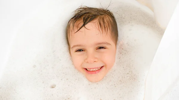 Retrato de niño alegre acostado en el baño con espuma de jabón y mirando en la cámara. Concepto de higiene infantil y atención sanitaria en el hogar. Niño divirtiéndose y jugando en casa. — Foto de Stock