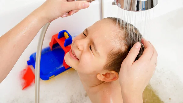 Niño pequeño cerrando los ojos mientras la madre se lava la cabeza con champú bajo la ducha en el baño. Concepto de higiene infantil y atención sanitaria en el hogar. — Foto de Stock