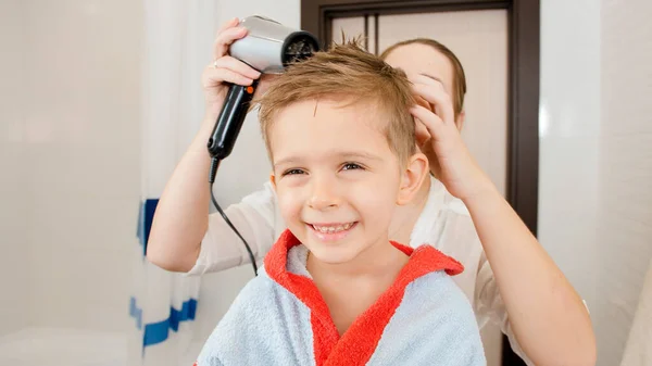 Счастливый улыбающийся маленький мальчик, смотрящий в зеркало в ванной, в то время как мама сушит его волосы феном. Концепция детской гигиены и здравоохранения на дому. Забота о родителях и детях — стоковое фото
