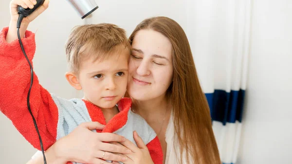 Äiti ja pieni poika kylpytakissa leikkii ja pitää hauskaa hiustenkuivaajalla kylpyhuoneessa. Käsitys lasten hygieniasta ja terveydenhoidosta kotona. Vanhemmat ja lapset hauskaa ja leikkii kotona — kuvapankkivalokuva