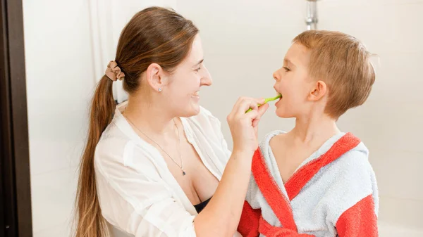 Портрет улыбающейся матери, чистящей зубы своего маленького сына в ванной. Концепция детской гигиены и здравоохранения на дому. Забота о родителях и детях. — стоковое фото