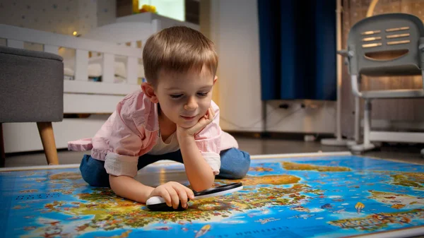 寝室の床に横になり、虫眼鏡を通して大きな世界地図を見ている小さな笑顔の少年。旅行、観光、子供教育の概念。子供の探検と発見. — ストック写真