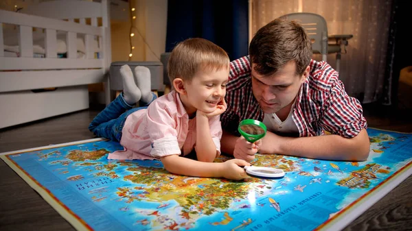 Divertido tiro de feliz padre msiling con su hijo pequeño explorando el mapa del mundo y jugando con lupas. Concepto de viajes, turismo y educación infantil. Exploración y descubrimiento infantil —  Fotos de Stock