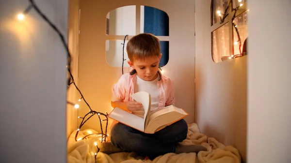 Cute little boy sitting in toy cardboard house at night and reading big fairy tale book. Concept of child education and reading in dark room — Stock Photo, Image