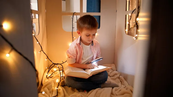 Bonito menino lendo livro com tocha em sua casa de papelão de brinquedo à noite. Conceito de educação infantil e leitura em sala escura — Fotografia de Stock