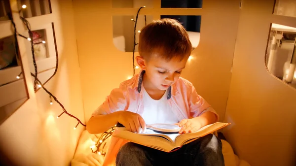 Petit garçon dans une maison de jouets avec des guirlandes légères lisant le livre la nuit. Concept d'éducation et de lecture des enfants en chambre noire — Photo