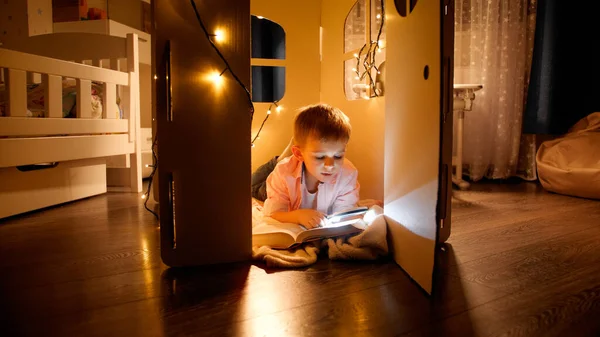 Feliz niño sonriente tumbado en el suelo en la casa de juguetes o tienda de campaña y leyendo con linterna por la noche. Concepto de educación y lectura infantil en cuarto oscuro — Foto de Stock