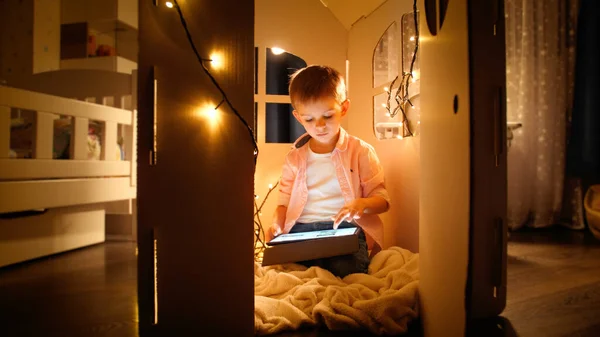 Kleine slimme jongen zittend in speelgoed kartonnen huis en met behulp van tablet computer. Begrip "kinderopvoeding" en "s nachts studeren — Stockfoto