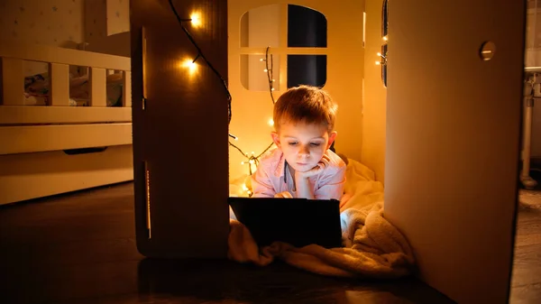 Kleine slimme jongen ligt in een klein speelgoedhuisje en uing tablet computer 's nachts. Begrip "kinderopvoeding" en "s nachts studeren — Stockfoto