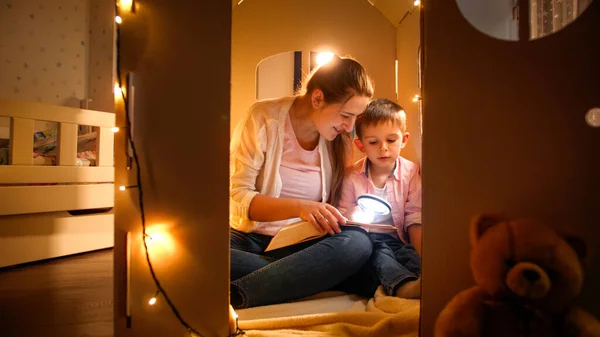 Glad leende pojke med ung mor sitter i tält eller leksakskartong hus och läsa bok med ficklampa på natten. Begreppet barnuppfostran och familj som har tid tillsammans på natten — Stockfoto