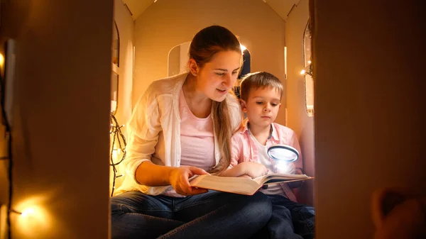 Portrait of smiling young mother with little boy reading book at night while playing in toy house. Concept of child education and family having time together at night — Stock Photo, Image