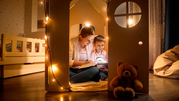 Młoda uśmiechnięta kobieta czytająca bajki swojemu synowi w nocy, siedząc na podłodze. Pojęcie wychowania dzieci i rodziny spędzających razem czas w nocy — Zdjęcie stockowe