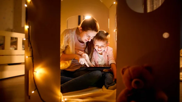 Glad skrattande pojke med ung mamma spelar spel och använder surfplatta dator på natten innan du går och lägger dig. Begreppet barnuppfostran och familj som har tid tillsammans på natten — Stockfoto