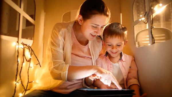 Portrait of happy smiling mother with little son playing and having fun with tablet computer in tent or small house at night. Concept of child education and family having time together at night — Stock Photo, Image