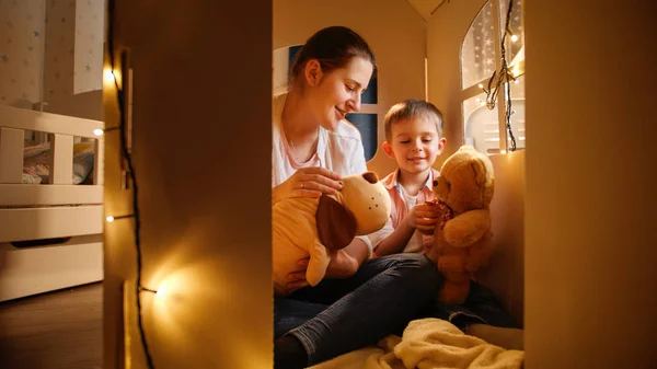 Menino bonito e jovem mãe jogando jogos com ursinhos de pelúcia de brinquedo à noite na tenda ou casa de brinquedo. Conceito de criança brincando e família tendo tempo juntos à noite. — Fotografia de Stock